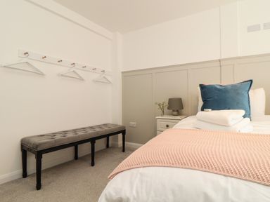 A bedroom with a bed with towels and cushions a bedside table with lamp and flowers a bench and coat hangers on the wall at The Old Post Office in Porthleven