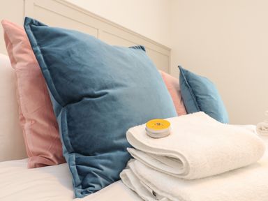 A close-up of a bed with blue and pink pillows and folded white towels with a small yellow tin on top at The Old Post Office in Porthleven