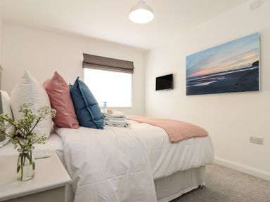 A bedroom with a bed with multiple pillows and a folded blanket on it a bedside table with a small vase of flowers a window with a blind a wall-mounted TV and a large painting at The Old Post Office in Porthleven