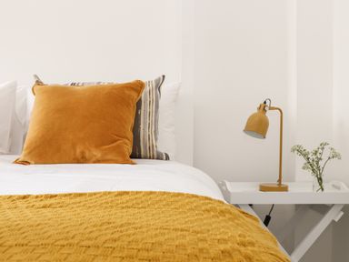 A bed with orange and striped pillows a yellow blanket a white bedside table with a yellow lamp and flowers at The Old Post Office in Porthleven