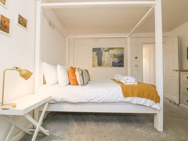 A bedroom with a white four-poster bed with pillows and towels a side table with a lamp and abstract art on the wall at The Old Post Office in Porthleven