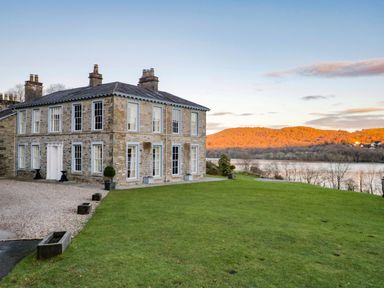 A stone house with multiple chimneys and white framed windows on a grassy lawn near a river with hills in the background at Silverholme Graythwaite near Hawkshead