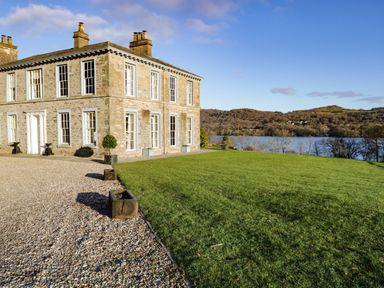 A stone house with large windows and chimneys next to a gravel path and a grassy lawn overlooking a lake and wooded hills at Silverholme in Graythwaite near Hawkshead