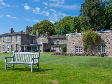 A stone house with multiple windows and chimneys with a wooden bench on a lawn at Silverholme Graythwaite near Hawkshead