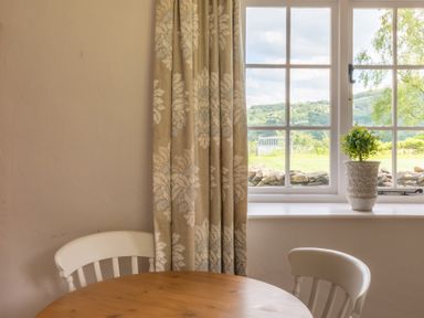 A round wooden table with two chairs near a window with patterned curtains and a potted plant on the window sill at Silverholme in Graythwaite near Hawkshead