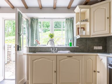 A kitchen with cream cabinets a sink under a window with green check curtains and an open door to the outside at Silverholme in Graythwaite near Hawkshead