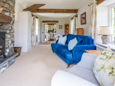 A living room with a blue sofa a fireplace exposed wooden beams and a doorway leading to a kitchen at Silverholme in Graythwaite near Hawkshead