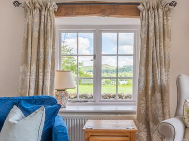 A window with floral curtains and a view of a garden and hills with a blue sofa and a grey armchair in a living room at Silverholme Graythwaite near Hawkshead