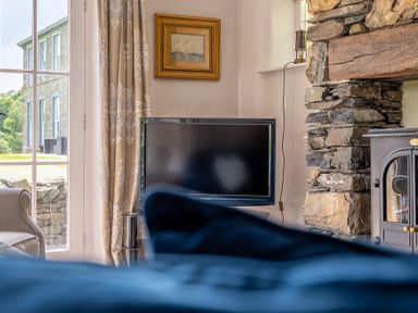 A living room corner with a television on a stand next to a stone fireplace and a window with curtains at Silverholme Graythwaite near Hawkshead