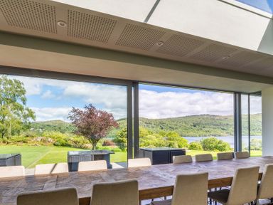 A dining room with a long wooden table and chairs facing large sliding windows overlooking a garden and hills at Silverholme in Graythwaite near Hawkshead