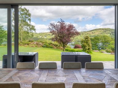 A dining table with chairs inside looking out through sliding glass doors to a patio with outdoor furniture and a garden with trees at Silverholme in Graythwaite near Hawkshead