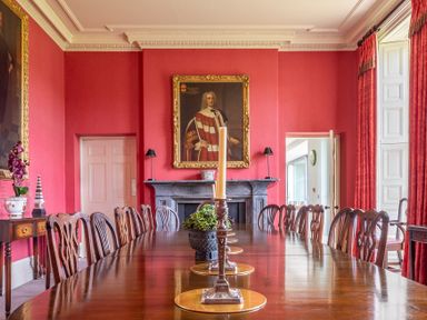 A dining room with a long wooden table and chairs red walls a fireplace and a portrait at Silverholme Graythwaite near Hawkshead