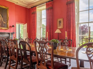 A dining room with a long wooden table candles and chairs by large windows with red curtains at Silverholme Graythwaite near Hawkshead