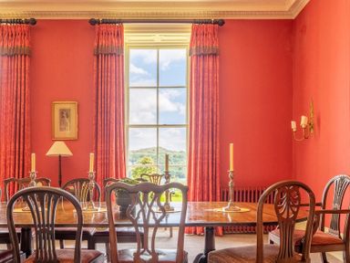 A dining room with wooden chairs and table with candles in front of a window with red curtains at Silverholme in Graythwaite near Hawkshead