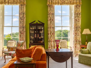 A sitting room with large windows floral curtains a china cabinet and a wooden table at Silverholme in Graythwaite near Hawkshead