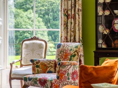 A room with a floral armchair a carved wooden chair a glass cabinet with china and floral curtains at Silverholme Graythwaite near Hawkshead