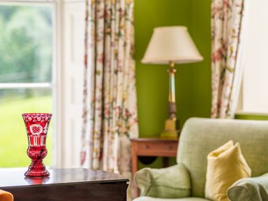 A red decorative glass vase on a wooden table with a green armchair and a lamp in a room with floral curtains and green walls at Silverholme Graythwaite near Hawkshead