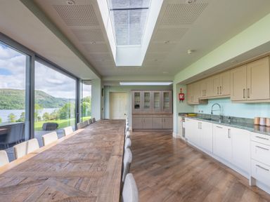 A kitchen with a long wooden dining table and chairs next to large windows overlooking a lake at Silverholme in Graythwaite near Hawkshead
