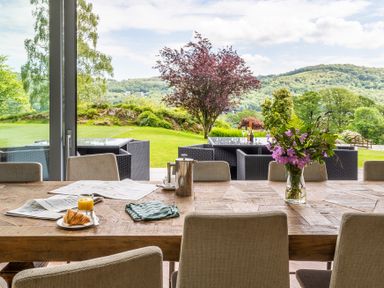 A dining table with chairs a vase of flowers and breakfast items overlooking a garden with outdoor seating and hills at Silverholme Graythwaite near Hawkshead