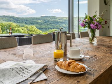 A wooden table with a croissant on a plate orange juice cup and newspaper by a window overlooking hills and a lake at Silverholme Graythwaite near Hawkshead