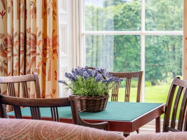 A dining table with a basket of lavender flowers surrounded by wooden chairs near a window with patterned curtains at Silverholme in Graythwaite near Hawkshead
