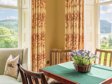 A corner dining area with wooden chairs and a green tablecloth on the table with a basket of flowers in front of windows with floral curtains at Silverholme Graythwaite near Hawkshead