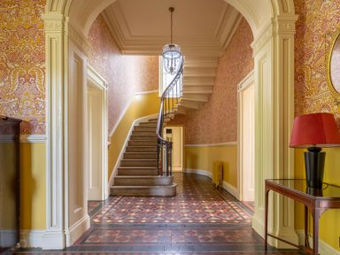 A hallway with patterned tiled floor a staircase with a black iron railing and a table with a red lamp at Silverholme Graythwaite near Hawkshead