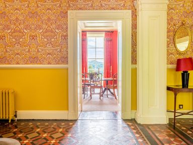An entryway with patterned floor tiles yellow wall paneling and ornate wallpaper leading to a dining room with wooden chairs and curtains at Silverholme in Graythwaite near Hawkshead