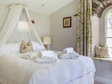 A bedroom with a bed covered in white linens and towels on it a lamp a window seat with floral cushions and curtains and a framed picture on the wall at Silverholme in Graythwaite near Hawkshead