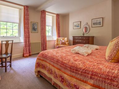 A bedroom with a bed covered in patterned bedspread two windows with curtains a wooden chair an armchair and chest of drawers with a mirror at Silverholme in Graythwaite near Hawkshead