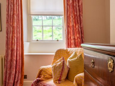 A corner of a room with a window covered by red patterned curtains a cushioned armchair with yellow and red patterned fabric and pillows and a wooden chest of drawers at Silverholme in Graythwaite near Hawkshead