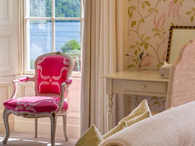 A room with a pink upholstered chair near a window and a white desk with flowers and a mirror at Silverholme Graythwaite near Hawkshead