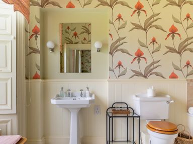 A bathroom with a pedestal sink a toilet with wooden seat a small black side table and floral wallpaper at Silverholme in Graythwaite near Hawkshead