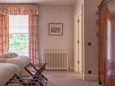 A bedroom with two beds a window with floral curtains a radiator a wooden wardrobe and a framed picture on the wall at Silverholme in Graythwaite near Hawkshead