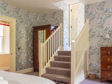 A bedroom area with floral wallpaper a wardrobe a chest of drawers stairs leading to a bathroom door at Silverholme in Graythwaite near Hawkshead