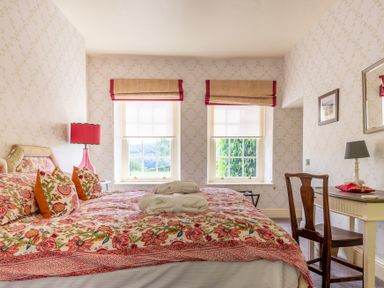 A bedroom with a bed covered in floral patterned bedding and pillows two windows with blinds a wooden chair and a dresser with a lamp at Silverholme Graythwaite near Hawkshead
