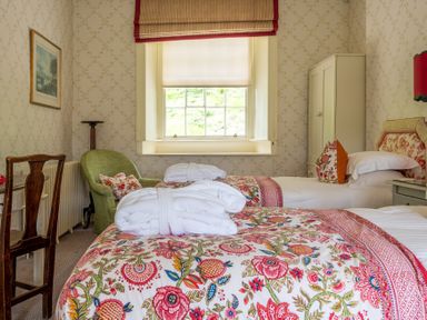 A bedroom with two single beds, floral bed covers, a green armchair, a wooden chair, a window with blinds and a white wardrobe at Silverholme Graythwaite near Hawkshead