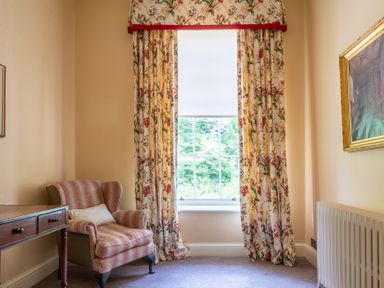 A corner of a room with a floral curtain on a window an armchair next to a wooden table and a radiator on the wall at Silverholme Graythwaite near Hawkshead