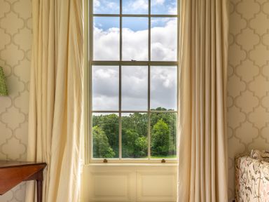 A large window with cream curtains showing green trees outside a room with patterned wallpaper and a wooden table