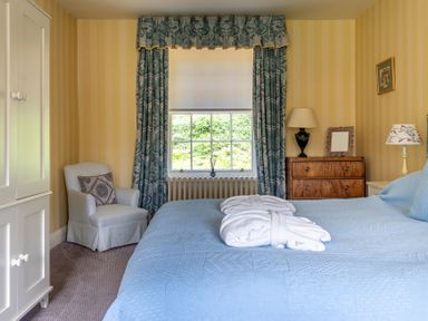 A bedroom with a blue bedspread two white robes a white armchair a wooden dresser and window with blue curtains at Silverholme Graythwaite near Hawkshead