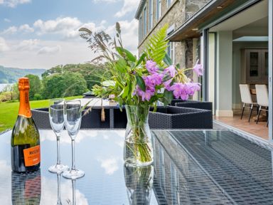 A glass table with a flower vase a bottle of prosecco and two glasses on a patio with outdoor chairs and a view of greenery at Silverholme Graythwaite near Hawkshead