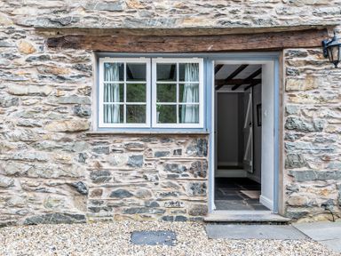 Stone exterior wall with a window and an open door showing a hallway with wooden beams on the ceiling at Silverholme in Graythwaite near Hawkshead
