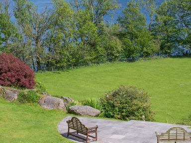 A garden with two wooden benches on a stone patio facing a grassy area with rocks bushes and trees near water at Silverholme in Graythwaite near Hawkshead