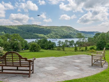 A patio with two wooden benches overlooking a lake and green hills with a plane in the sky at Silverholme in Graythwaite near Hawkshead