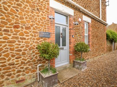 Stone exterior wall with a grey door named Troon Cottage and two potted plants on a gravel path at Troon Cottage Hunstanton