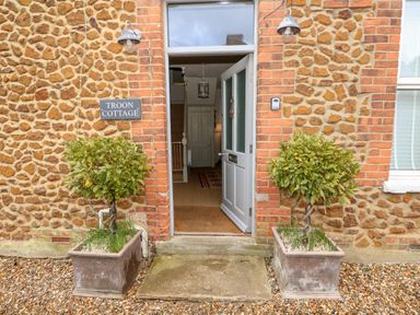 An open front door of a stone and brick cottage with two potted plants on either side at Troon Cottage Hunstanton