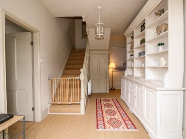 A hallway with a staircase and baby gate a white cabinet with shelves and a patterned rug at Troon Cottage in Hunstanton