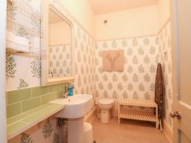 A bathroom with a pedestal sink and mirror a wall-mounted wire shelf a toilet a wooden bench and an ironing board at Troon Cottage in Hunstanton