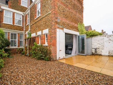 A gravel and paved courtyard with plants near a stone and brick building with windows and an open glass door at Troon Cottage in Hunstanton