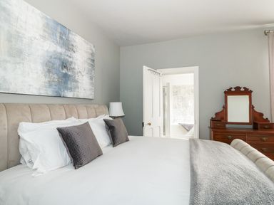 A bedroom with a bed with pillows a lamp a dresser with a mirror and an open door leading to a bathroom at Troon Cottage Hunstanton